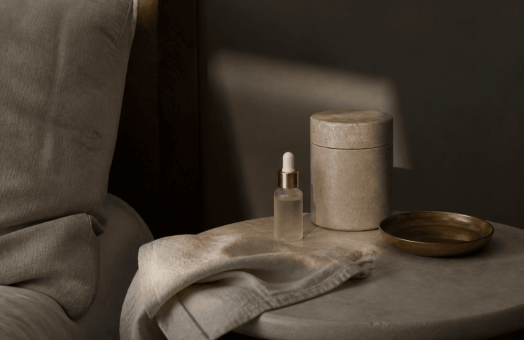 Lumière tamisée du soir sur une table de chevet sobre, un flacon discret et du linge clair — ambiance repos, beauty sleep.